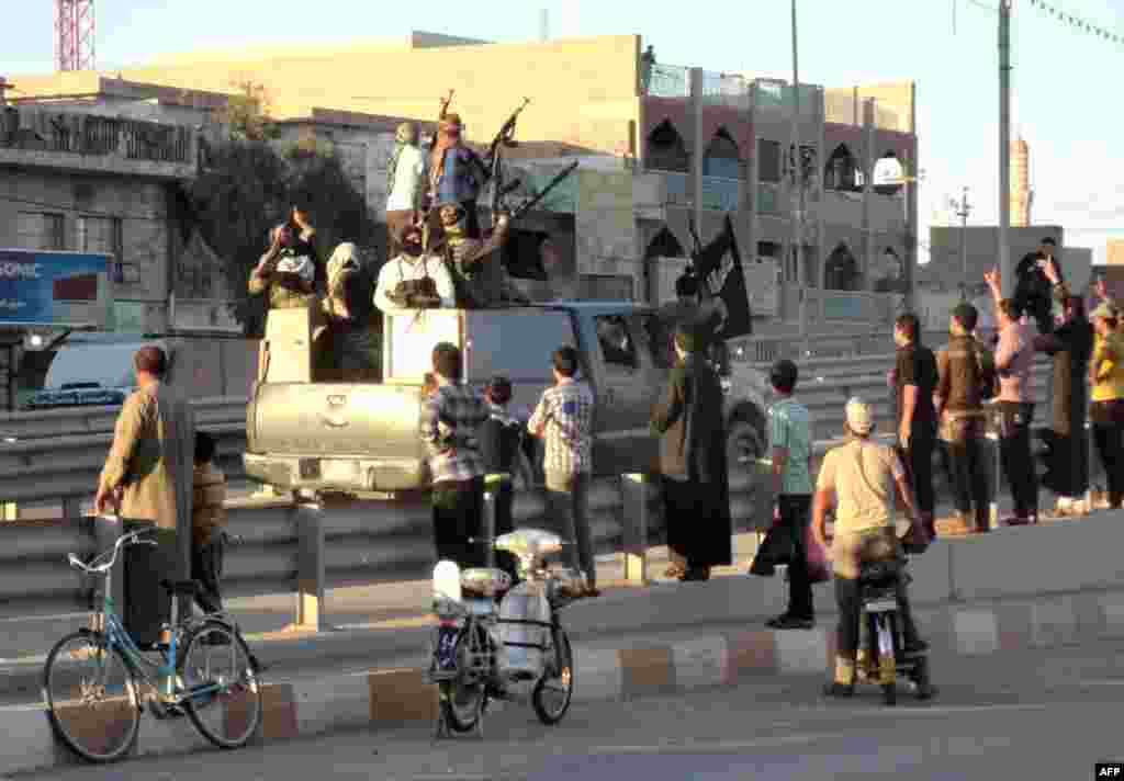 In Fallujah, life goes on. Local Iraqis stand watching an armored vehicle carrying militants holding an Islamic jihadist flag.